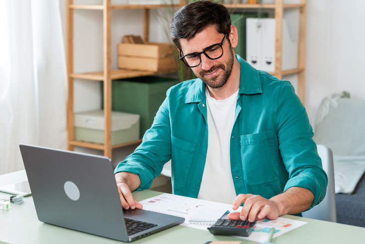 Smiling man working on laptop with calculator in home office