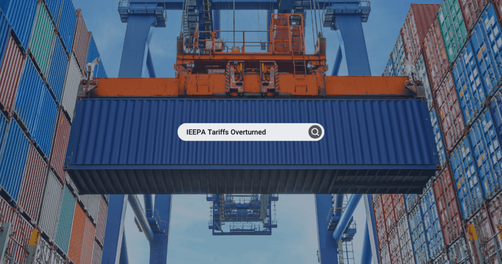 A shipping container being hoisted by a blue gantry crane in a busy port terminal, with stacks of colorful red, blue, and green containers in the background. Centered on the image is a search bar containing the text "IEEPA Tariffs Overturned" next to a magnifying glass icon.