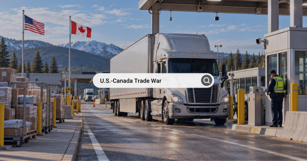 Truck crossing border at U.S.-Canada trade checkpoint with flags and mountains.